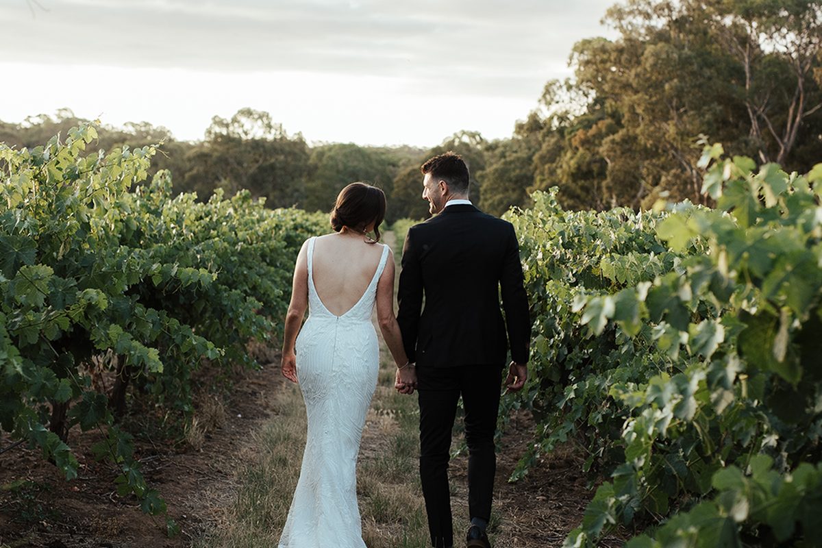 A bride and groom walk hand in hand down a grassy row between green vines at sunset.