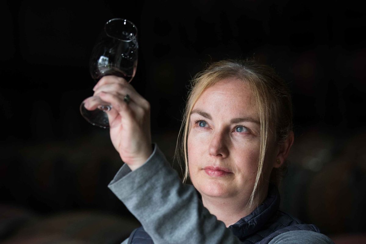 Winemaker Brooke Blair holds a glass of red wine up to the light in a dim barrel hall.