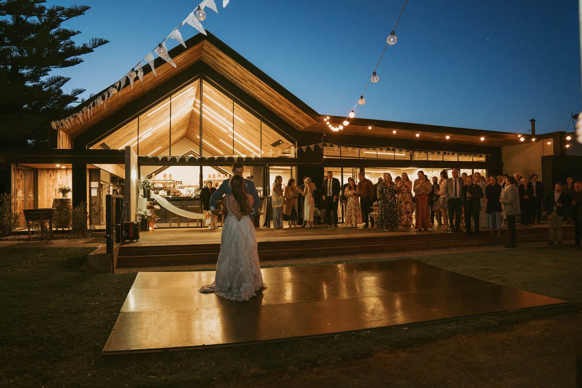 A couple share their first dance on a dance floor laid on the lawn at dusk, guests watching from the lit cellar door deck.