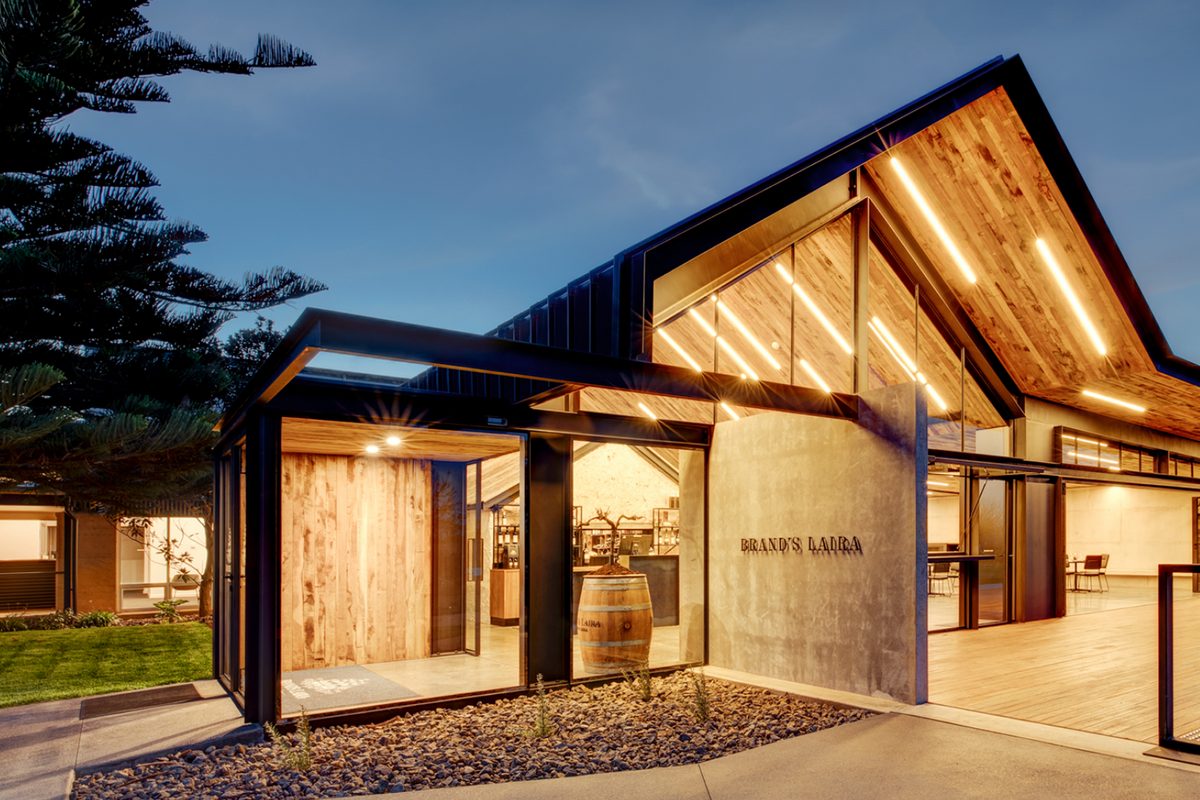 The Brand's Laira cellar door at dusk, its timber-lined gable glowing through floor-to-ceiling glass.