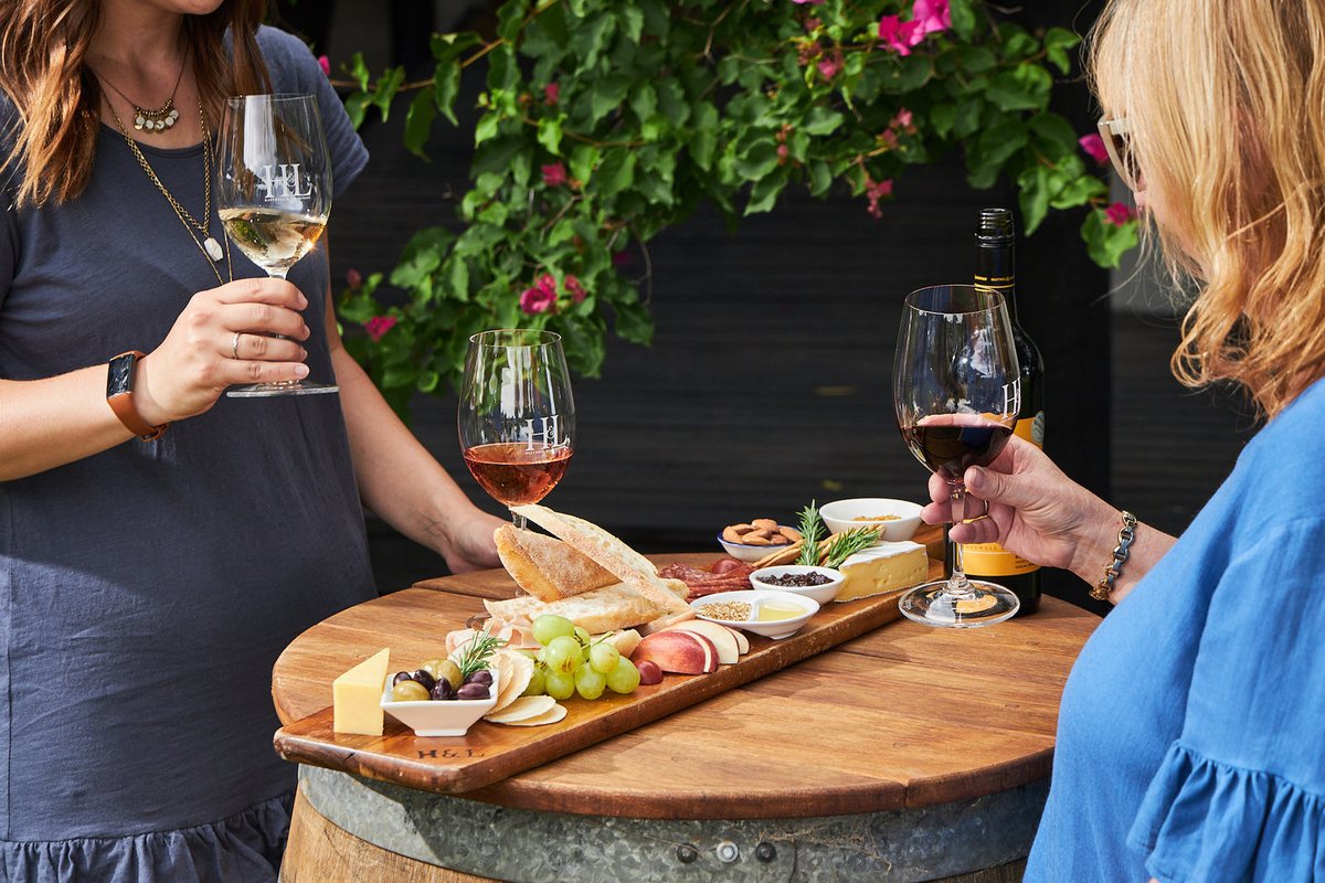 Two friends raise glasses of white and rosé over a grazing board of bread, cheese, grapes and olives set on a wine barrel.