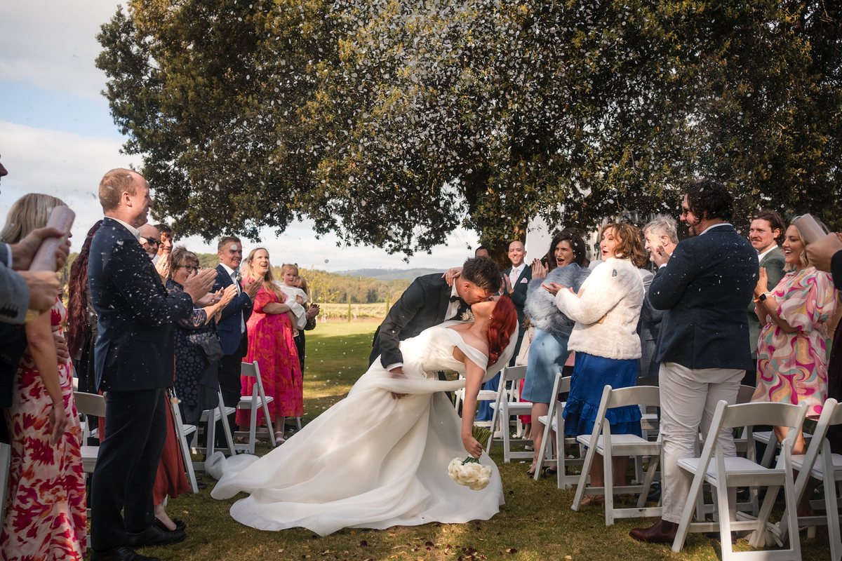 Guests throw confetti as a groom dips his bride for a kiss at the end of the aisle, beneath a spreading tree beside the vineyard.